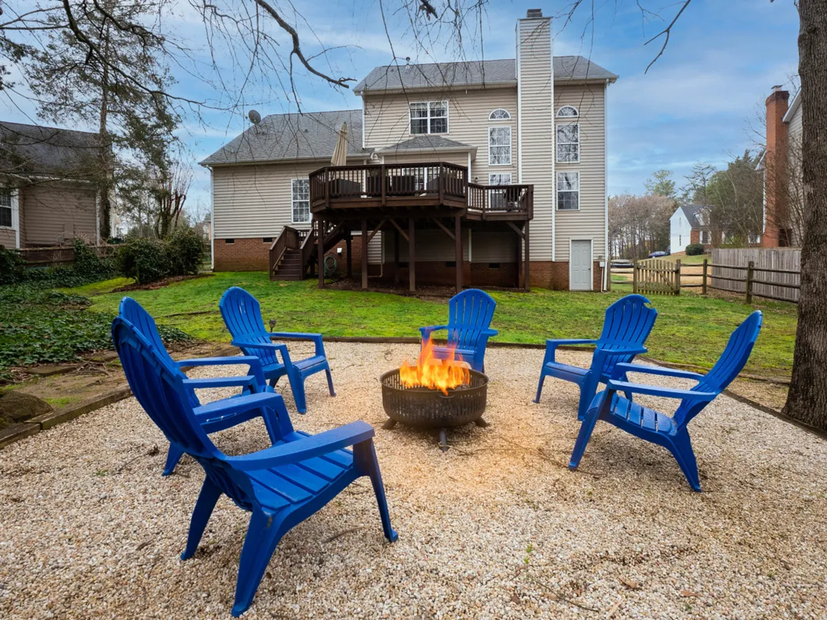 This exterior image showcases a spacious wooden deck with a Charbroil grill ready for barbecuing, a prominent wood-burning firepit surrounded by blue chairs, and the lush, wooded backdrop of the backyard. The deck and firepit area are set up for social gatherings, with inviting seating and a communal atmosphere.