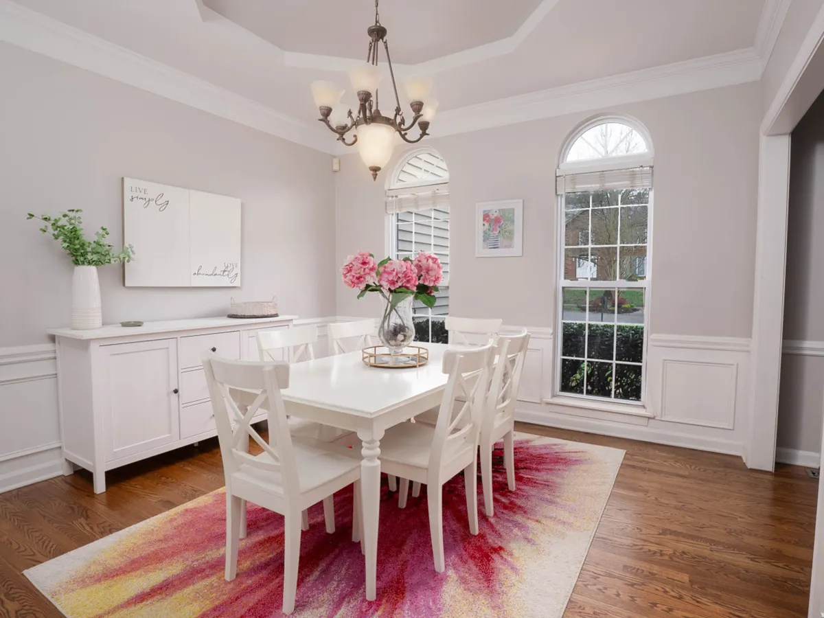 A formal dining area with a large table set for eight, accented by a statement chandelier, floral centerpiece, and a patterned rug. The setting feels both elegant and welcoming, highlighting ample seating for shared meals or celebrations.