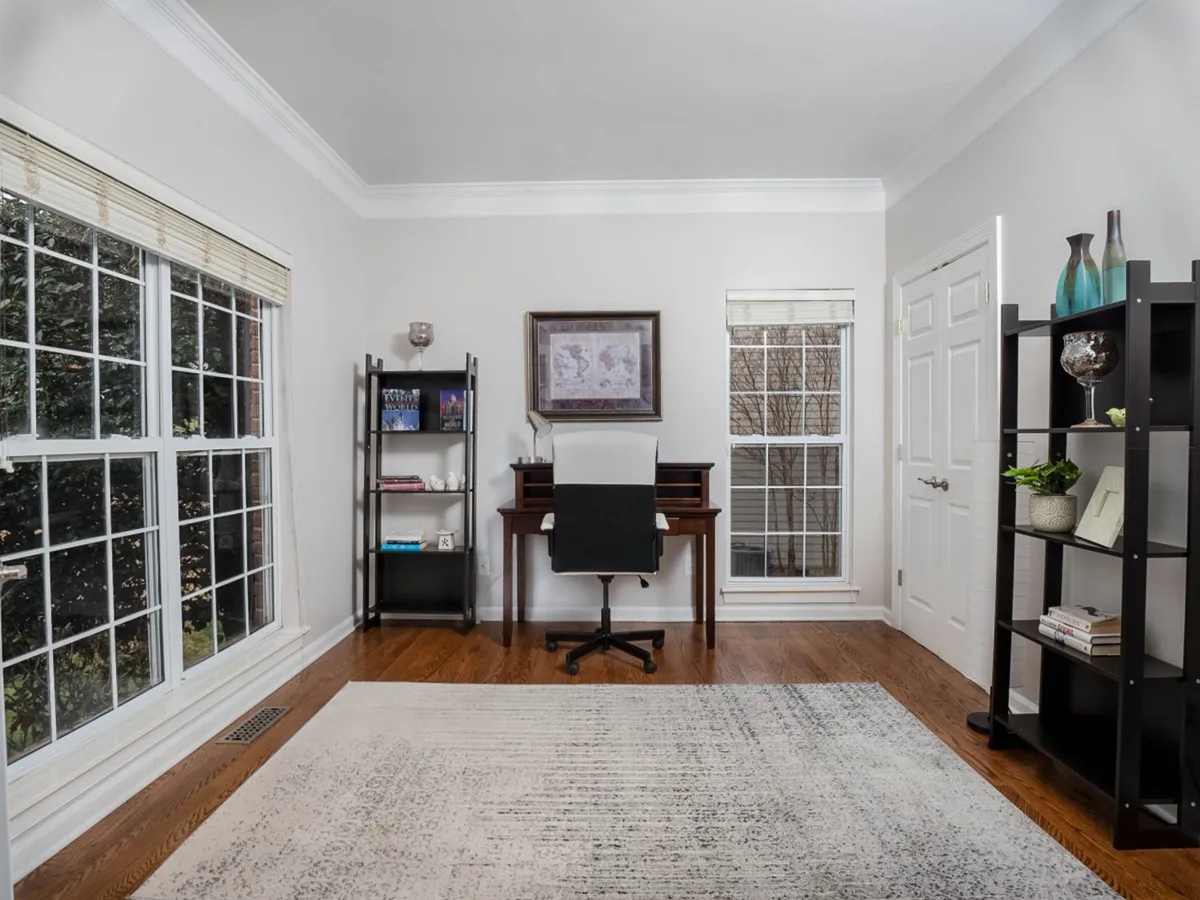 A sunlit office space featuring a desk with a comfortable chair and a bookshelf, all framed by large windows that invite in abundant natural light. The serene setup is ideal for focused work, reading, or study, and exudes a productive yet calming atmosphere.