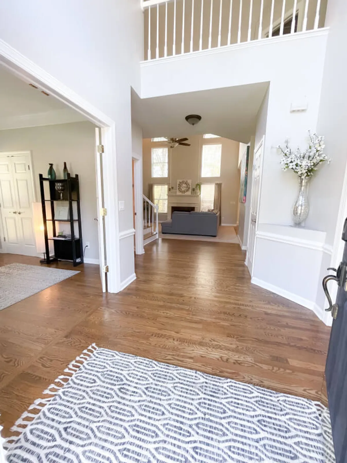 The image shows the inviting entryway of the property, including an entrance rug, a staircase leading to the upper floor, a sofa visible in the living room, and a decorative vase. The space is bright and open, hinting at a warm welcome for guests arriving for work or leisure.