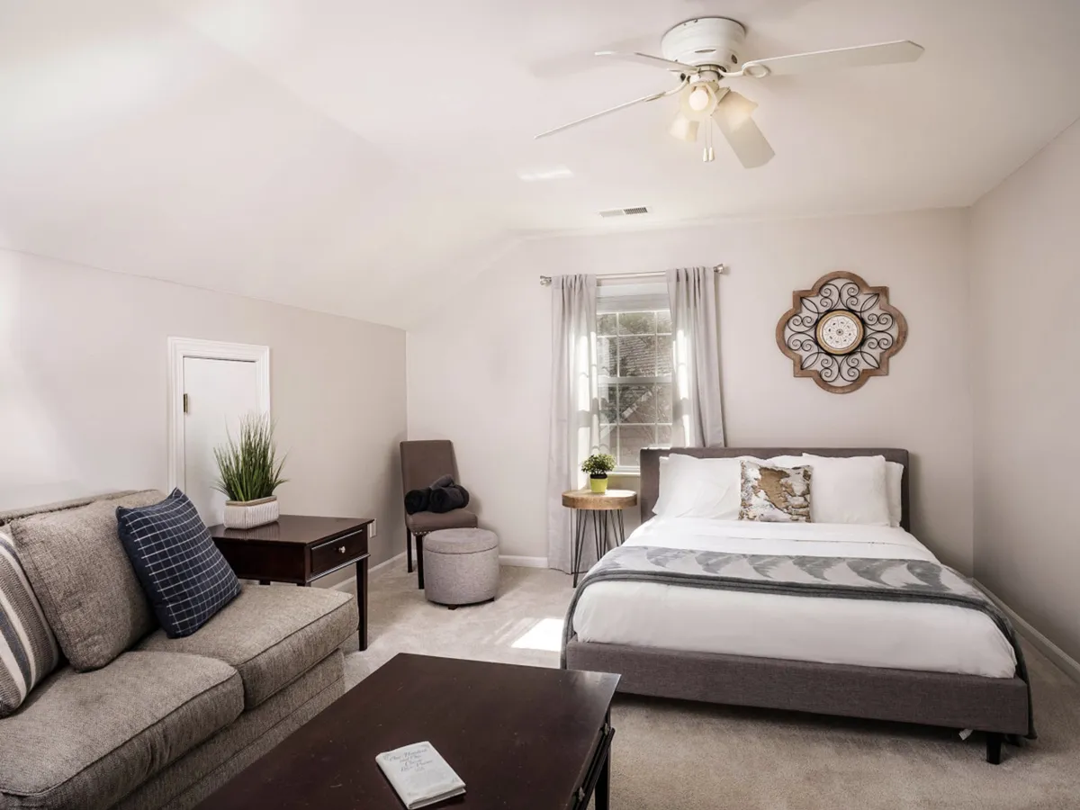 A comforting and tranquil bedroom and sitting area, featuring a bed with sunlight streaming in, a cozy sofa, decorative wall clock, and soft curtains. The setting evokes morning rituals and peaceful relaxation, ideal for couples seeking intimacy and comfort.
