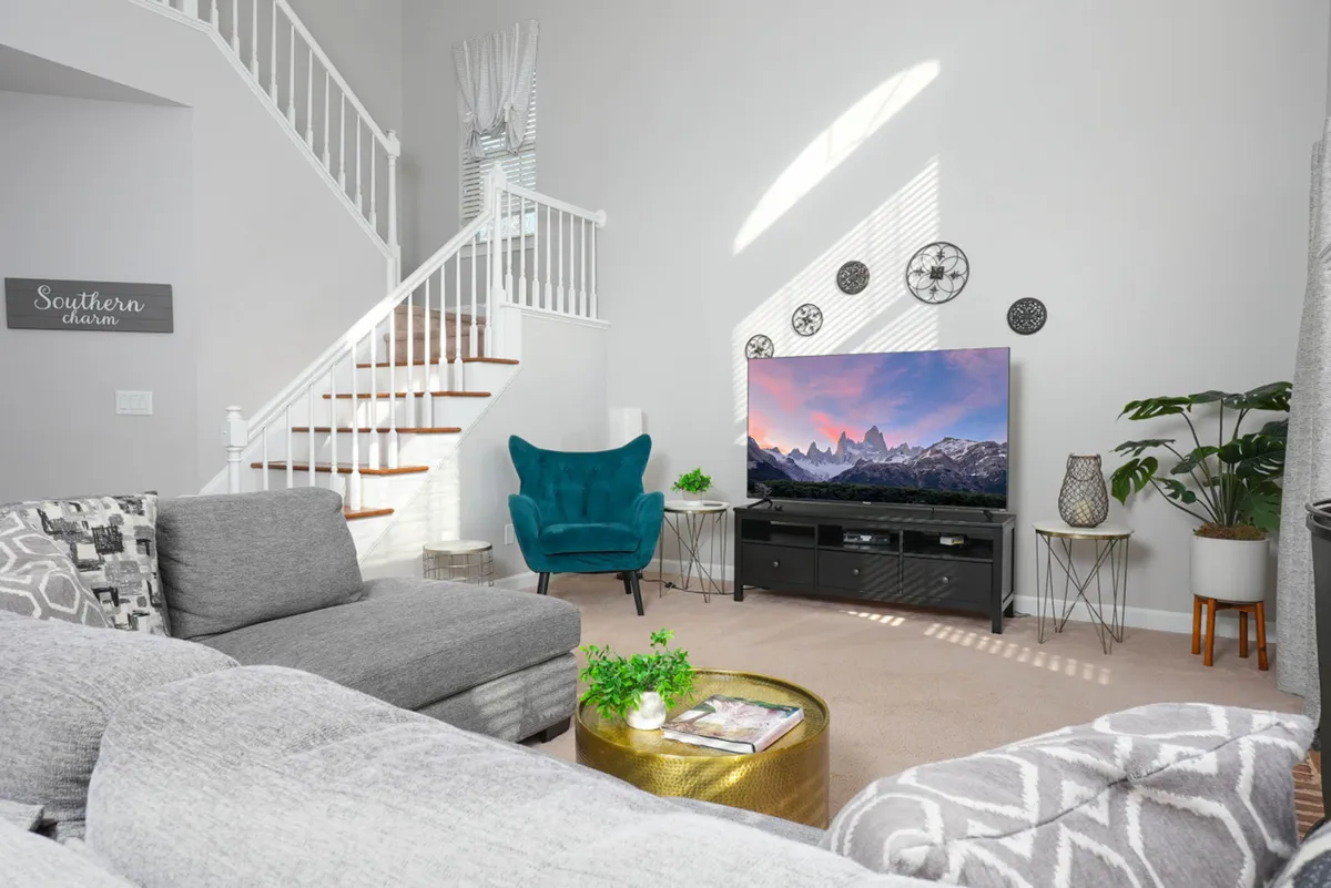 A spacious living room with a large gray sectional sofa, a big TV displaying a scenic mountain landscape, a teal accent chair, decorative wall plates, and a green potted plant. The setting is bright and designed for comfort, with ample space for group gatherings and relaxation.