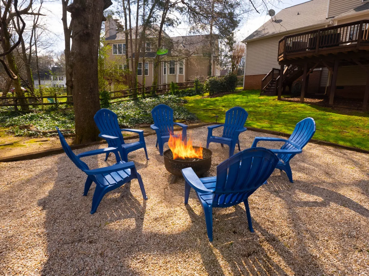 An intimate setting with a firepit encircled by blue chairs beneath the shade of surrounding trees. The area is designed for cozy evenings, storytelling, and toasting marshmallows under the stars, reinforcing the family-friendly, communal vibe.