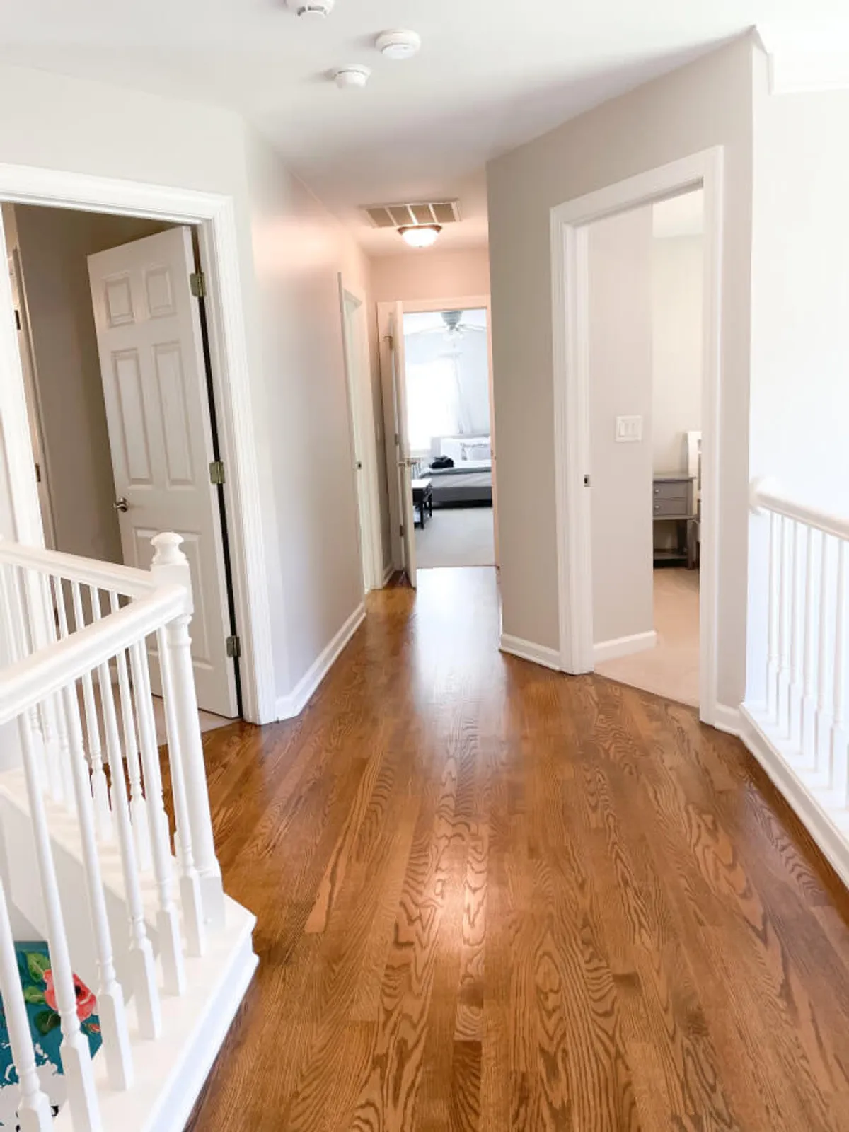 A bright hallway with wooden flooring, white railings, and open doors, filled with morning sunlight. People are seen walking and engaging in light conversation, suggesting an environment that is both social and serene.