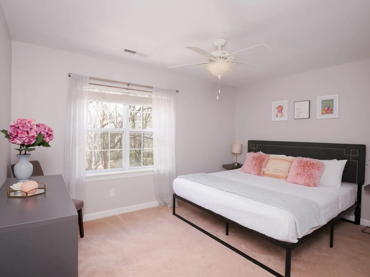 A tranquil, cozy bedroom with decorative pillows on the bed, sheer curtains that let natural light pour in, floral decor, and artwork. The scene suggests relaxing moments, enjoying coffee by the window, and quiet time for couples to reconnect.