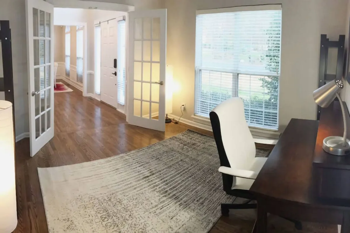 A thoughtfully arranged workspace featuring a desk, ergonomic chair, cozy rug, and ample windows for natural light, accented by stylish light fixtures. The room feels both functional and inviting, ideal for productive work sessions or quiet reading.