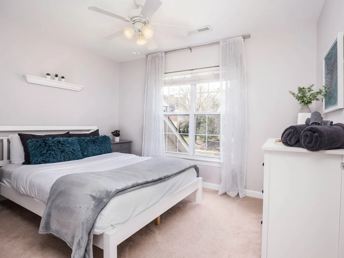 A serene bedroom with a comfortable bed adorned with decorative pillows, a large window with sheer curtains letting in natural light, and a white dresser. The ambiance is cozy and restful, suitable for couples or families. The space looks flexible and homey, ideal for any guest arrangement.