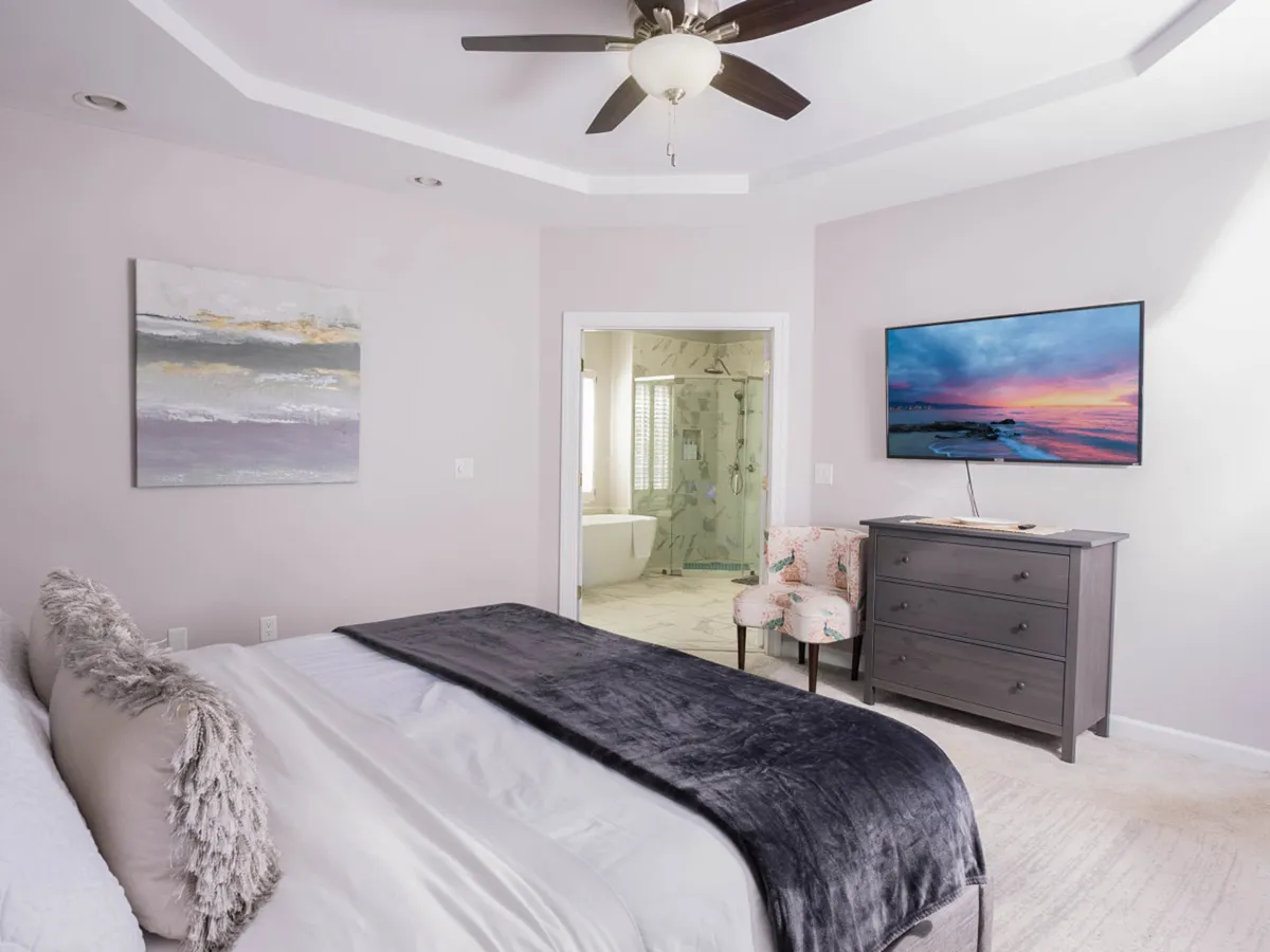 This image presents a serene bedroom with a plush bed, soft bedding, wall art, and a television. A dressing table with chairs sits near a doorway leading to an inviting bathroom, promising both comfort and convenience. The overall feeling is cozy and restful, with natural light creating a tranquil atmosphere.